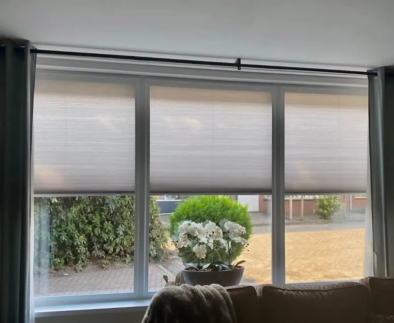 Lichtdoorlatende honingraat plisségordijnen in grijs in een woonkamer met drie ramen en uitzicht op de tuin