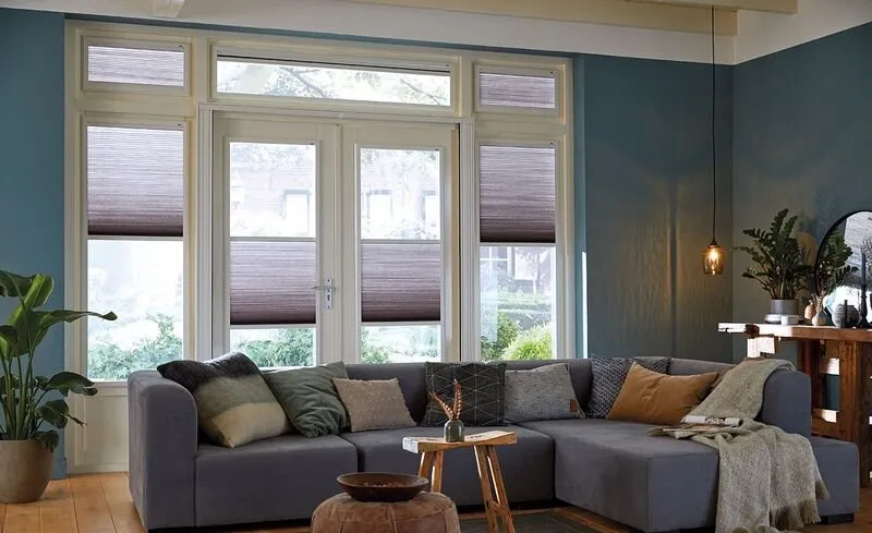 Lichtdoorlatende plissegordijnen in taupe in een sfeervolle woonkamer met grote ramen en blauwe muren